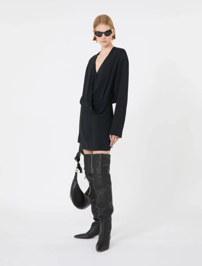 Model in an all-black outfit: draped V-neck blouse with a short skirt, knee-high black boots, black sunglasses, and a crescent-shaped handbag