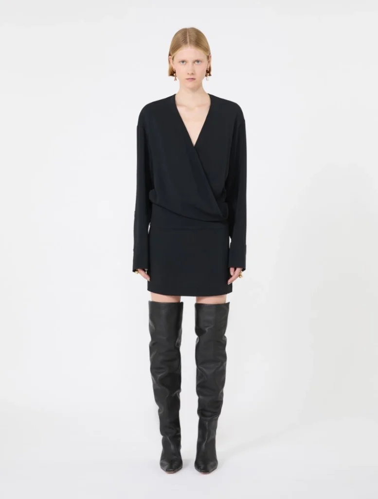 Front view of a model in a black V-neck wrap blouse, matching mini skirt, and tall black leather boots.
