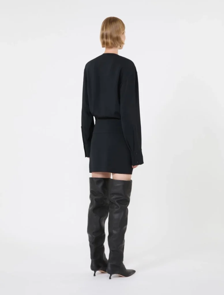 Back view of a person wearing a black long-sleeve blouse tucked into a black mini skirt, paired with over-the-knee black leather boots.