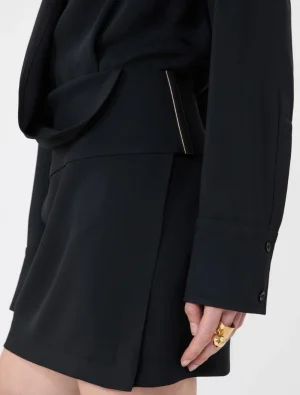 Close-up of a black tailored blazer with a waist belt, featuring a slim metallic rectangular accessory peeking from the belt and a hand with gold rings nearby.