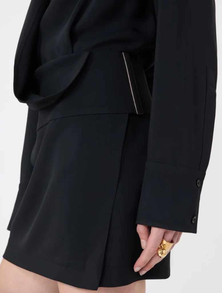 Close-up of a black tailored blazer with a waist belt, featuring a slim metallic rectangular accessory peeking from the belt and a hand with gold rings nearby.