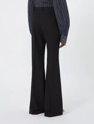 Back view of a person wearing black wide-leg trousers and a dark pinstriped shirt with cuffed sleeves, standing against a light background.