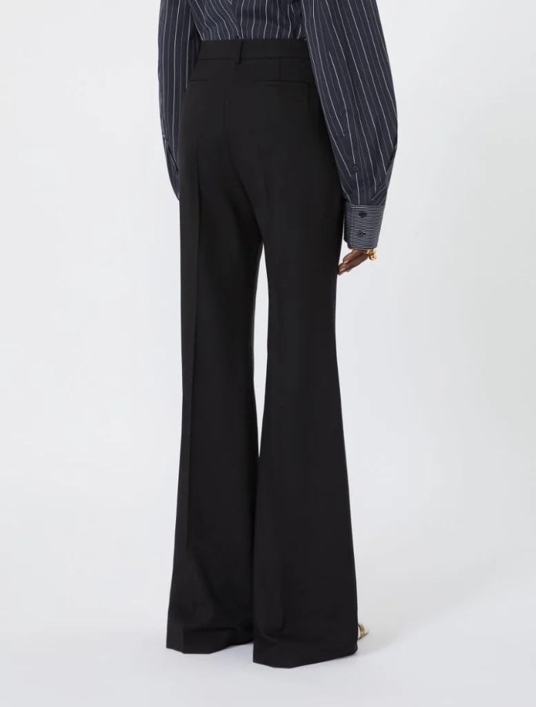 Back view of a person wearing black wide-leg trousers and a dark pinstriped shirt with cuffed sleeves, standing against a light background.