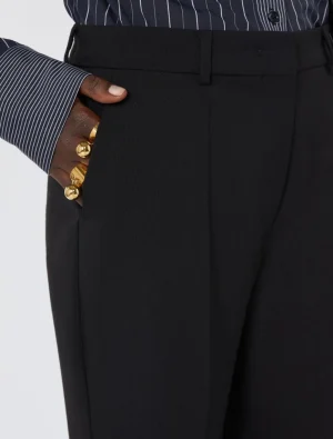 Close-up of a person wearing black tailored trousers with a hand in the front pocket; visible gold rings and a striped shirt cuff.