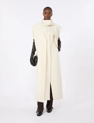 Model wearing a long cream coat with high neck, black gloves, brown pants, and black boots in a minimalist studio.