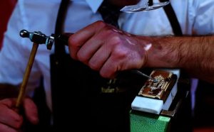 Close-up of a craftsman hammering and chiseling a decorative gold plate clamped in a bench vise at a workbench.