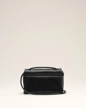 Black leather handbag with a top handle and long shoulder strap against a pale beige backdrop