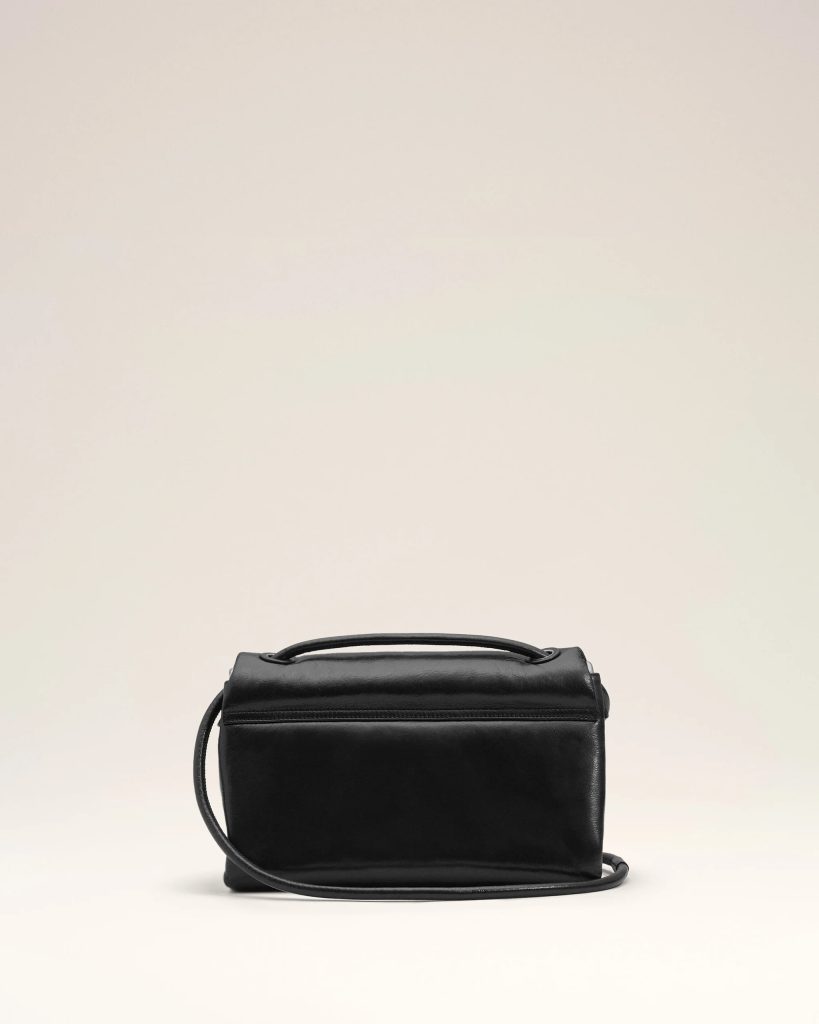 Black leather handbag with a top handle and long shoulder strap against a pale beige backdrop
