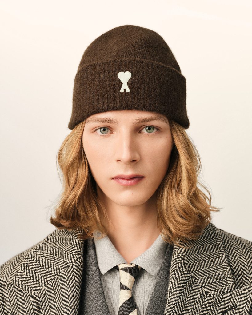 Person wearing a dark brown knit beanie with a small white heart and A emblem, paired with a gray blazer and striped tie.