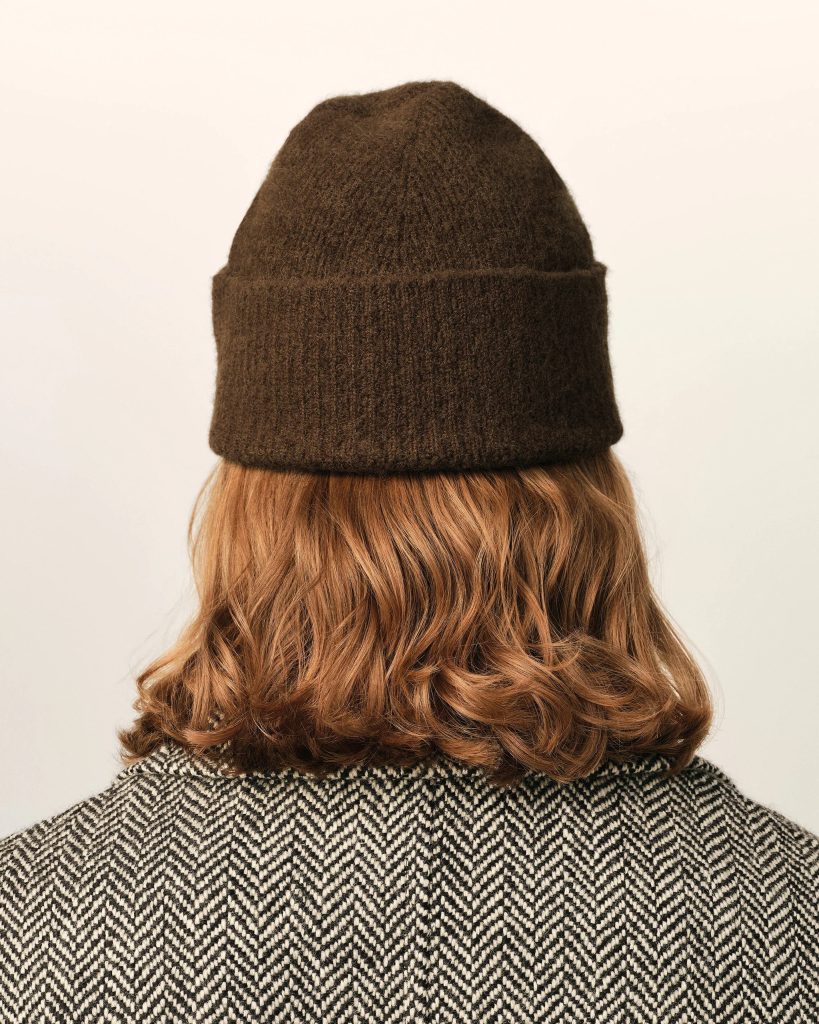 Back view of a person wearing a dark brown knit beanie and a black-and-white herringbone coat, blonde hair peeking from below the hat