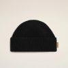 Black knit beanie hat laid flat on a pale background, showing folded cuff and small brown tag on the side.