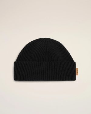 Black knit beanie hat laid flat on a pale background, showing folded cuff and small brown tag on the side.