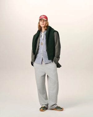 Full-body portrait of a person in a striped shirt, gray coat, and a green sweater draped over the shoulders, wearing a red cap and green sneakers.