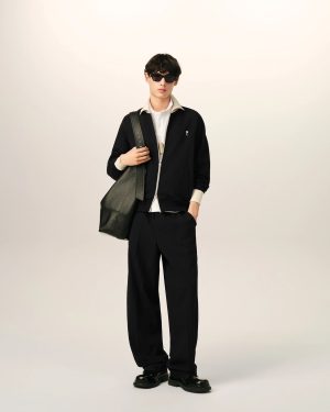 Person wearing a black jacket and pants with a white-collar shirt, sunglasses, and a large shoulder bag.