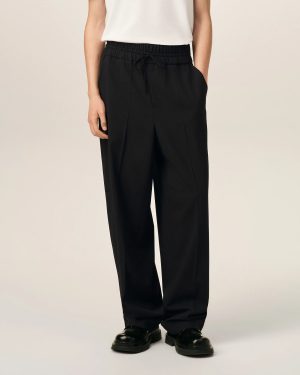 Person wearing black drawstring trousers and black shoes, standing against a light backdrop. Option: Focus on clothing: black pants with elastic waistband and drawstring, paired with black shoes.