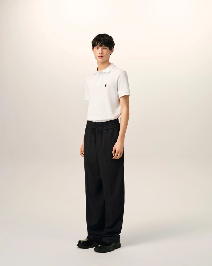Full-body portrait of a person wearing a white polo shirt, black drawstring pants, and black shoes, standing against a light backdrop