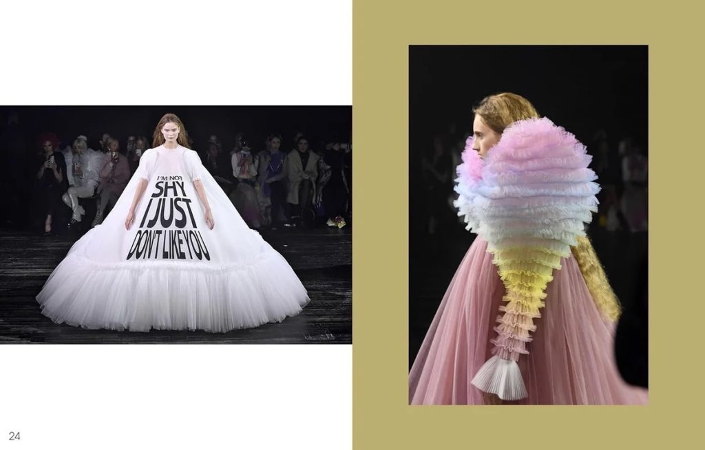 Runway model in a voluminous white gown with bold black text reading “I'M NOT SHY I JUST DON'T LIKE YOU.” visually connected to a fashion show audience in the background.