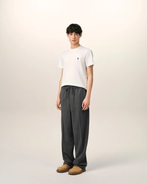 Full-body portrait of a young man standing against a pale background, wearing a white T-shirt, dark gray wide-leg pants, and tan shoes, facing the camera.