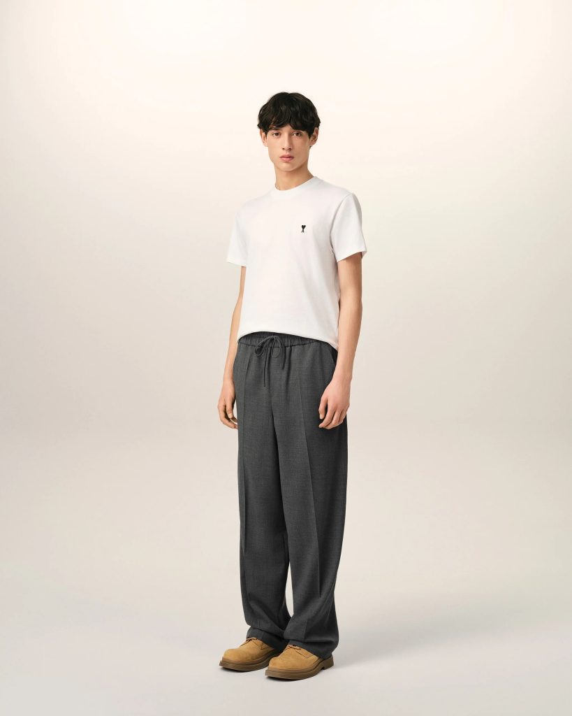 Full-body portrait of a young man standing against a pale background, wearing a white T-shirt, dark gray wide-leg pants, and tan shoes, facing the camera.