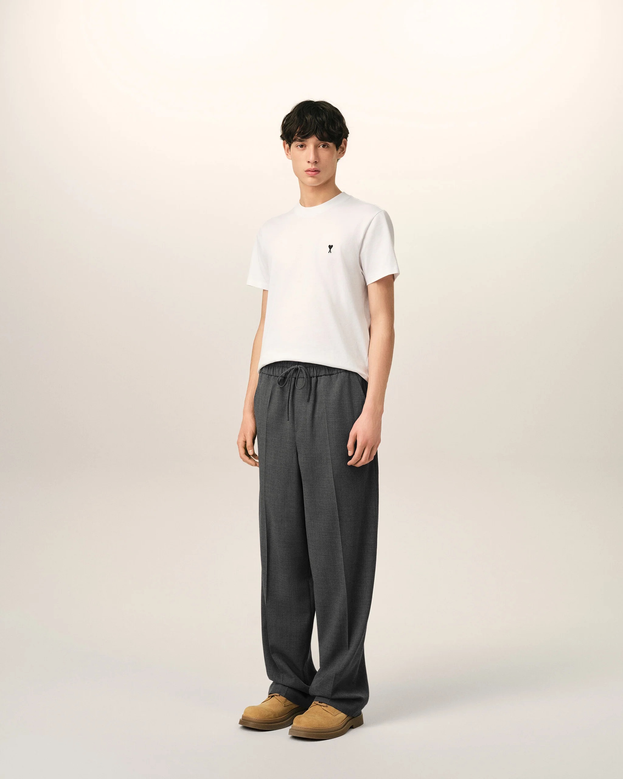 Image 143 - New Trend Full-body portrait of a young man standing against a pale background, wearing a white T-shirt, dark gray wide-leg pants, and tan shoes, facing the camera.