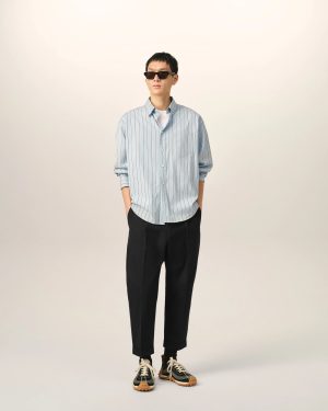 Person standing against a light neutral backdrop, wearing a light blue striped button-down shirt, black pants, and sneakers with sunglasses for a casual, fashion-focused look.
