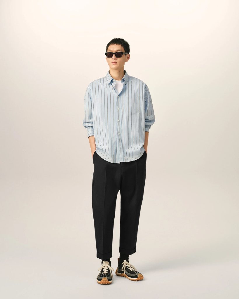 Person standing against a light neutral backdrop, wearing a light blue striped button-down shirt, black pants, and sneakers with sunglasses for a casual, fashion-focused look.