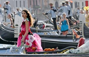 A woman in a bright pink floral gown stands in a gondola during a Venice parade, other gondolas and costumed performers in background