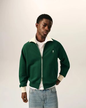 Man wearing a dark green zip-up jacket with a cream collar, white shirt underneath, and light denim jeans, posing against a pale background.