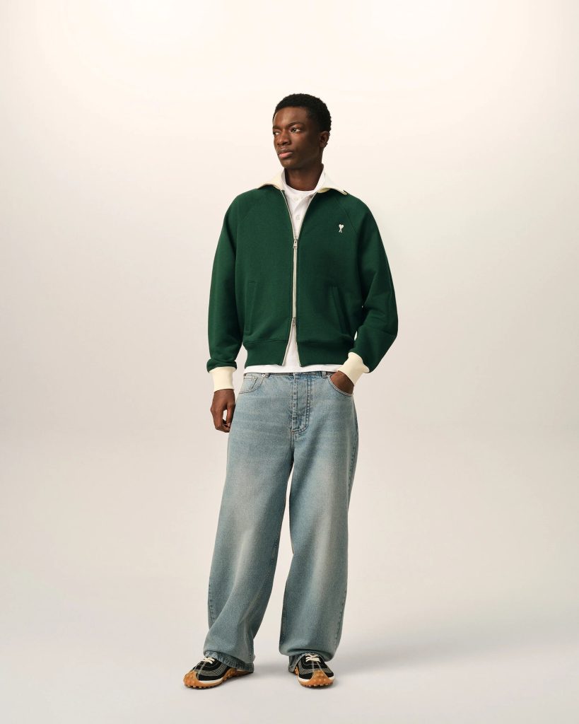 Male model wearing a dark green zip-up jacket over a white shirt, light blue jeans, and sneakers, standing in a neutral studio.