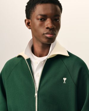 Close-up of a person wearing a dark green zip-front jacket over a white shirt, with a small white heart-shaped patch on the left chest against a beige background.