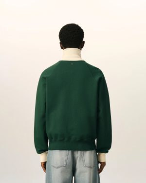 Person viewed from behind wearing a dark green sweater over a cream turtleneck and light-wash jeans against a pale background.