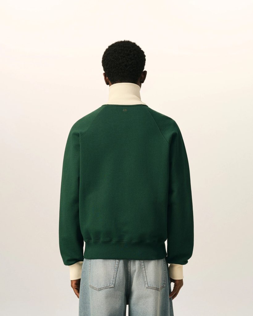 Person viewed from behind wearing a dark green sweater over a cream turtleneck and light-wash jeans against a pale background.