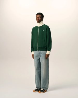 Full-body portrait of a man in a green zip-up jacket with a cream collar, light jeans, standing against a pale backdrop.