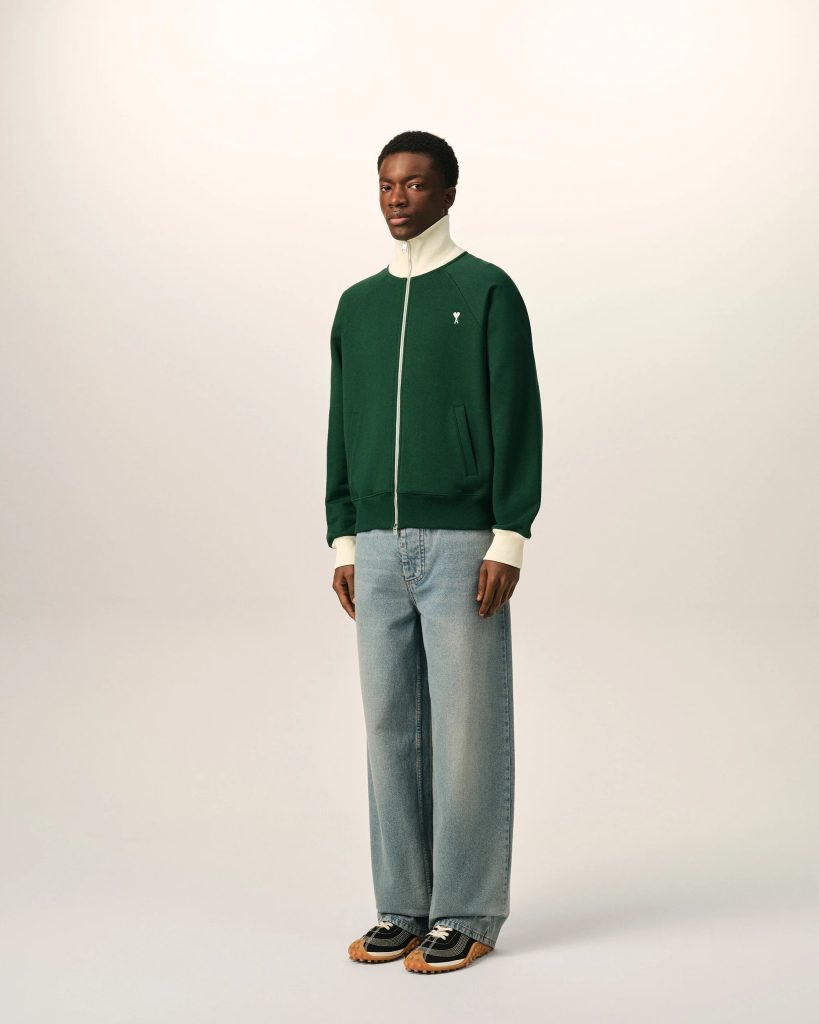 Full-body portrait of a man in a green zip-up jacket with a cream collar, light jeans, standing against a pale backdrop.