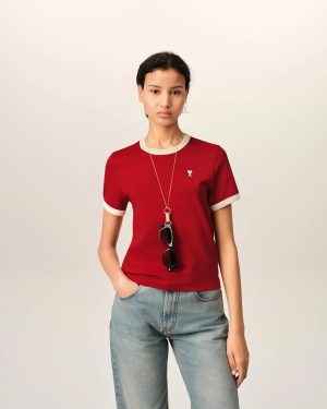 Person wearing a red top with beige trim and jeans, sunglasses hanging from the neckline, standing against a plain light background.