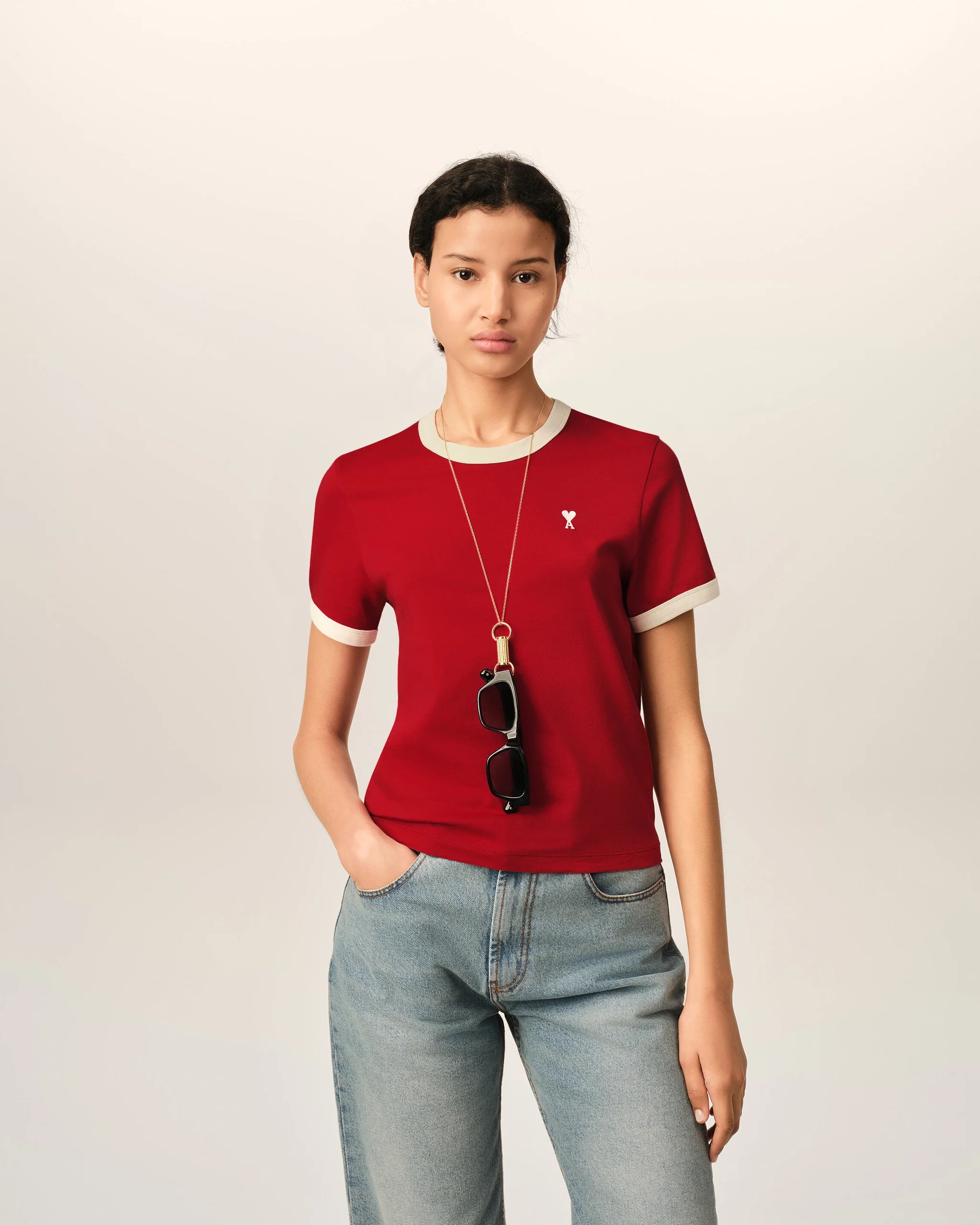 Image 159 - New Trend Person wearing a red top with beige trim and jeans, sunglasses hanging from the neckline, standing against a plain light background.