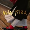 Gold 'New York' text overlaying a person in a black coat, gray skirt, red socks, and black loafers on stone steps.