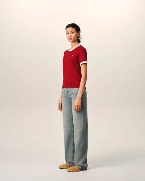 Full-body portrait of a young woman facing the camera, wearing a red short-sleeve shirt with white cuffs and collar, light blue jeans, and tan loafers against a neutral backdrop.