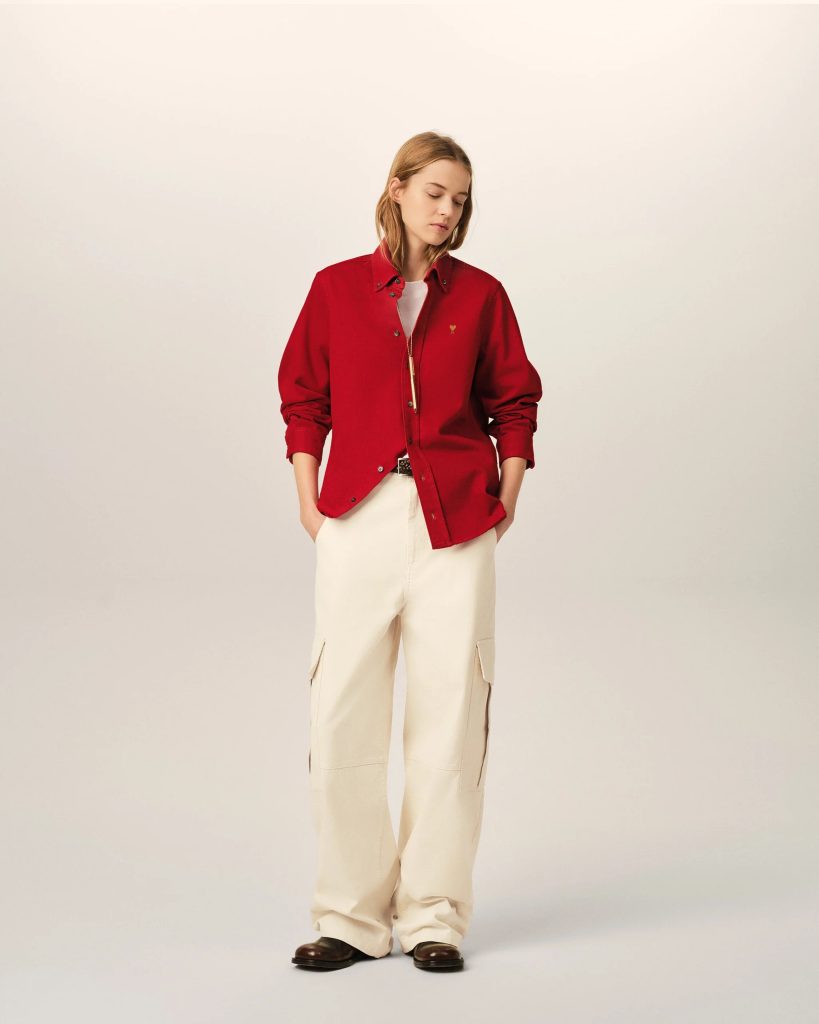 Full-body portrait of a woman wearing a red button-down shirt over a white top, cream cargo pants, and brown shoes, against a light backdrop.