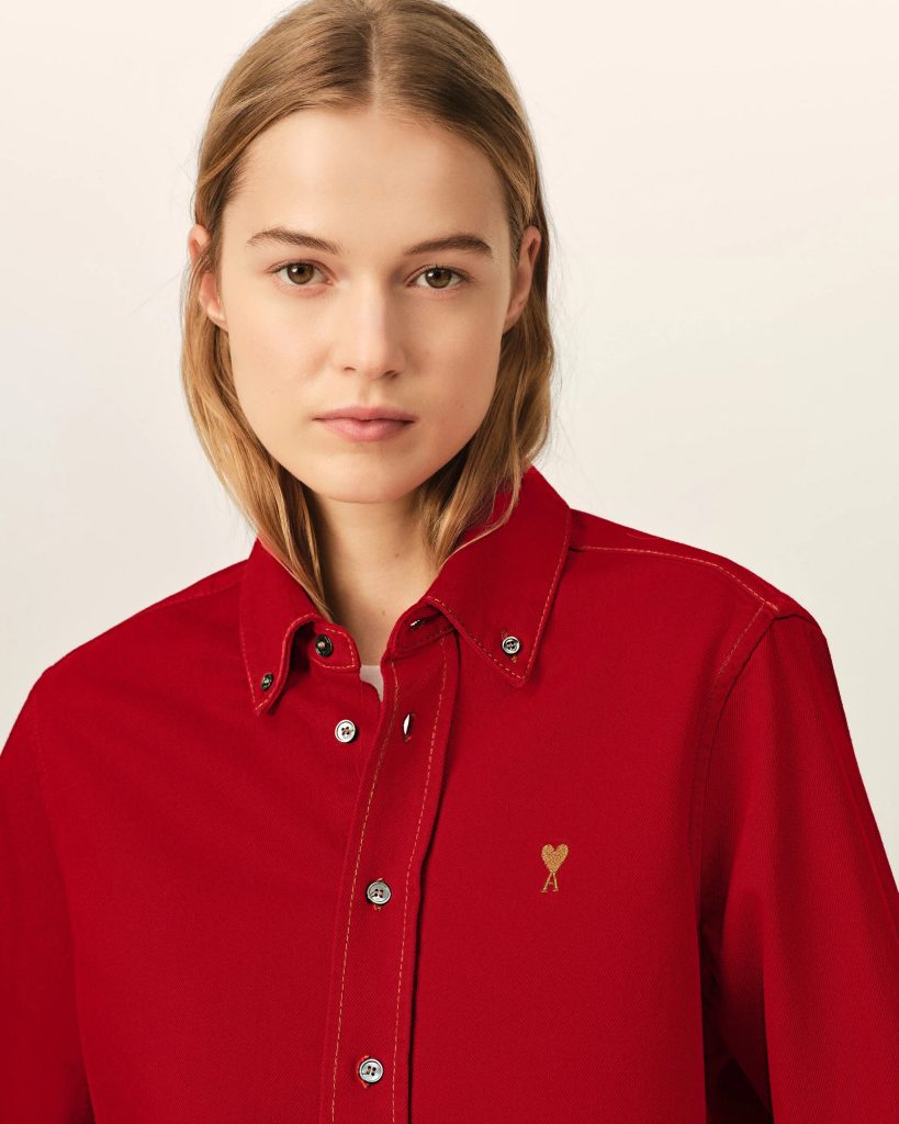 Close-up of a young woman with light brown hair wearing a red button-down shirt, neutral expression, and a small gold heart emblem on the left chest.