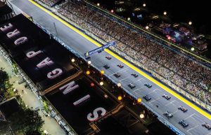 Aerial view of a nighttime Formula 1 race start at the Singapore Grand Prix with cars lined up on the grid and a packed grandstand.