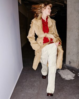 Woman wearing a tan faux fur coat over a red blouse and beige cargo pants, walking through a concrete backstage hallway with her hair swinging as she smiles.