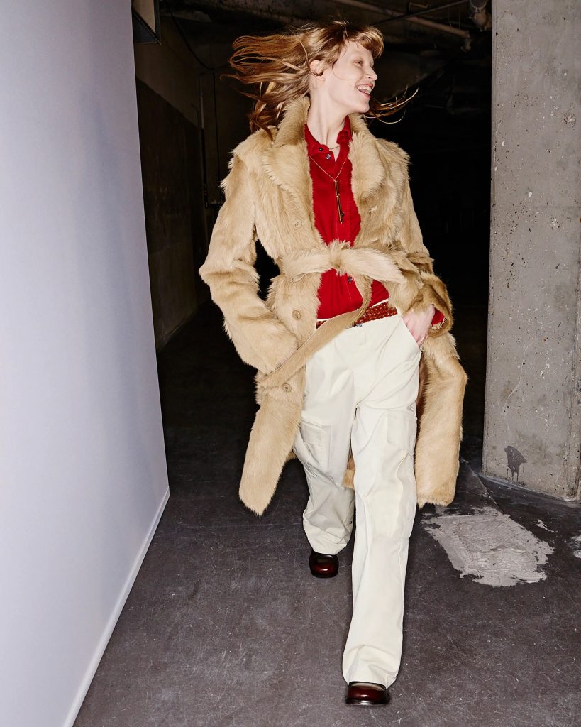 Woman wearing a tan faux fur coat over a red blouse and beige cargo pants, walking through a concrete backstage hallway with her hair swinging as she smiles.