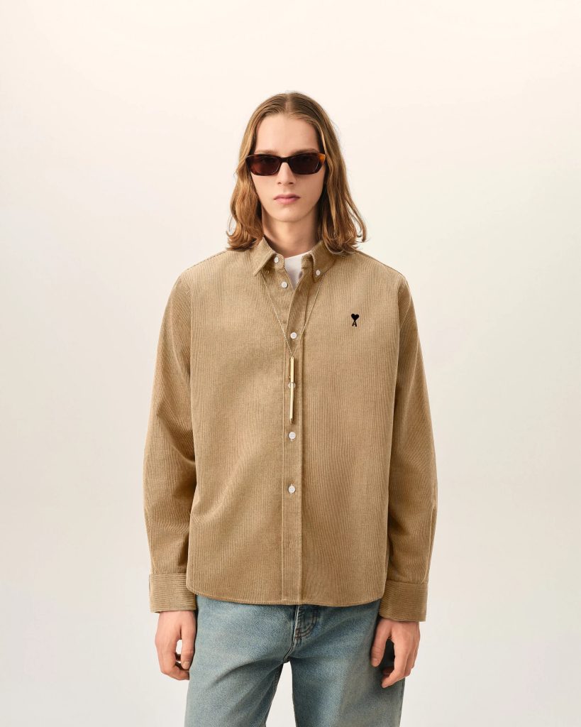 Person wearing a tan corduroy shirt with sunglasses and a long necklace, standing against a neutral wall.