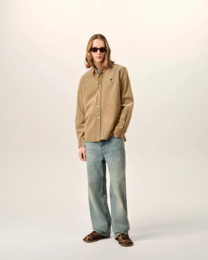 Person standing against a light neutral background wearing a tan button-down shirt, blue jeans, leopard-print shoes, and dark sunglasses.