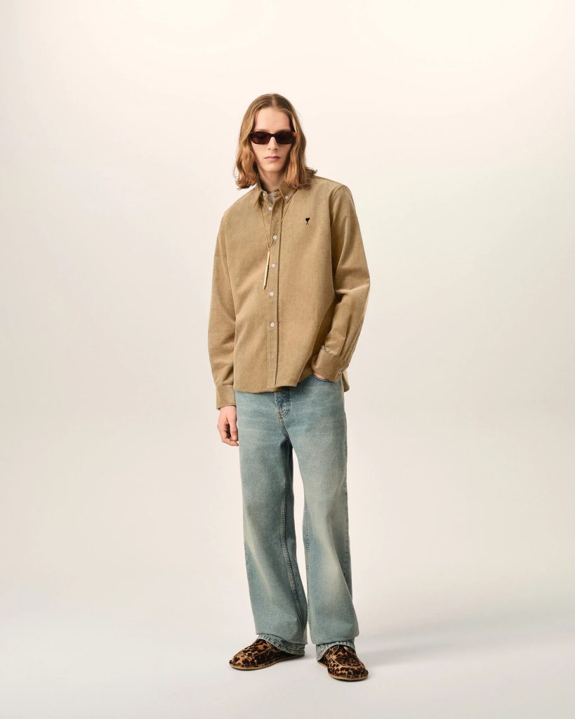 Person standing against a light neutral background wearing a tan button-down shirt, blue jeans, leopard-print shoes, and dark sunglasses.