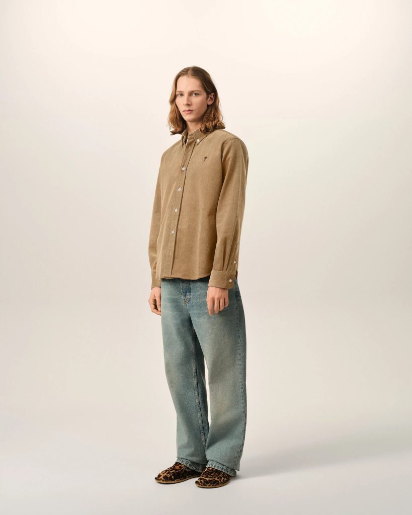 Full-body portrait of a person wearing a tan button-down shirt, blue jeans, and leopard-print shoes against a light backdrop (neutral studio setting).