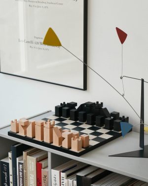 Wooden chess-like pieces (light and dark) arranged on a black-and-white checkered board on a desk, with a contemporary wire sculpture featuring colored shapes extending across the board and wall.