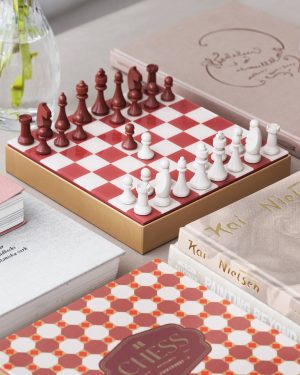 Red and white chess pieces arranged on a red-and-white checkered chessboard inside a box, with a glass vase in the background.