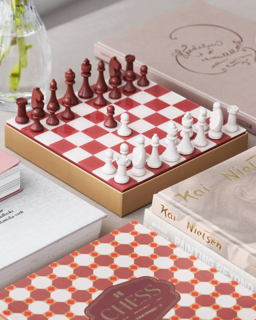 Red and white chess pieces arranged on a red-and-white checkered chessboard inside a box, with a glass vase in the background.